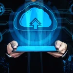 Businessman holding a tablet with a glowing blue cloud upload icon, surrounded by digital cloud computing and network technology symbols on a dark background.